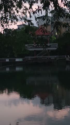 Pavilion with red tiled roof by reflective pond at dusk
A traditional-style pavilion with a red tiled roof stands on columns at the edge of a calm pond, connected by a walkway; lush trees surround the