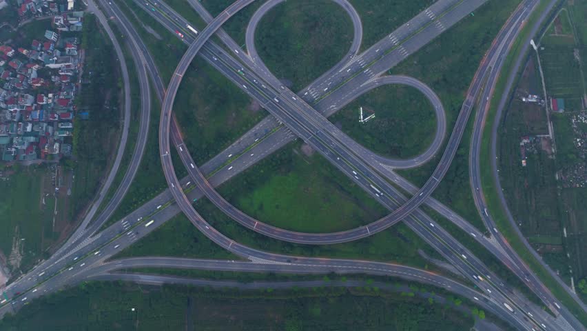An overhead hyperlapse drone shot of a complex highway interchange in Hanoi, Vietnam, with a cloverleaf design and light traffic.