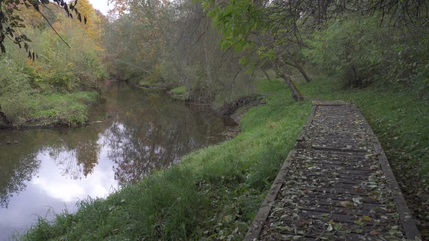 An old boardwalk winds beside a calm river, lined with golden foliage and fallen leaves, guiding the way through a peaceful autumn forest trail.