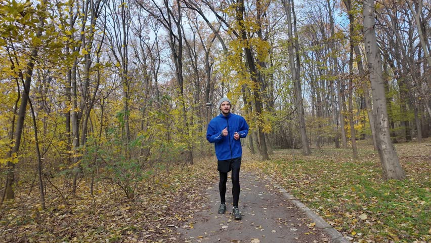 Man jogging in autumn forest wearing blue jacket and beanie, training on a leaf-covered path. Horizontal 4K footage.