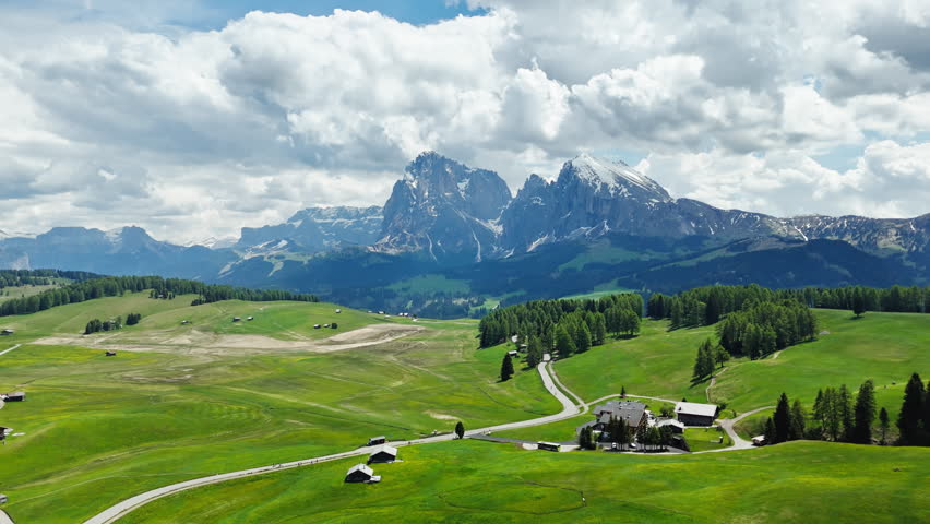 Curving farm tracks across alpine meadows with the Sassolungo group in background, aerial view. Rural mountain terrain in the Dolomites