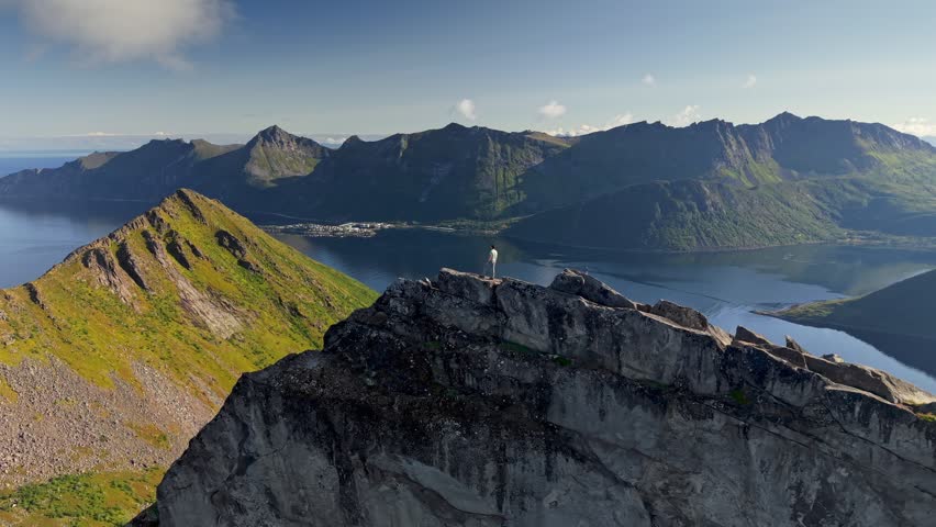 Hiker on rocky peak overlooking calm waters and majestic landscapes Senja island, Norway. Climber celebrating climbing top of Segla mountain by raising his arms 