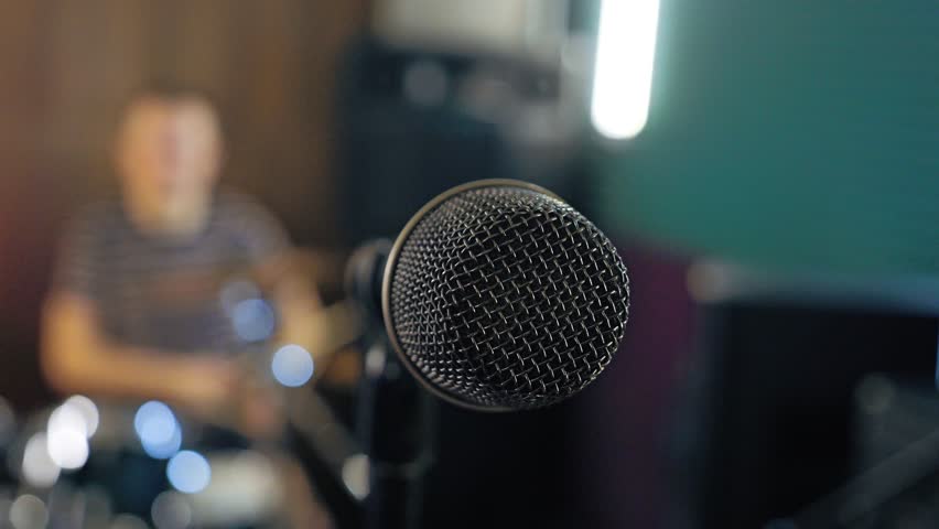 Close-up of microphone against blurred background, drummer playing drums. Camera moves around microphone from left to right