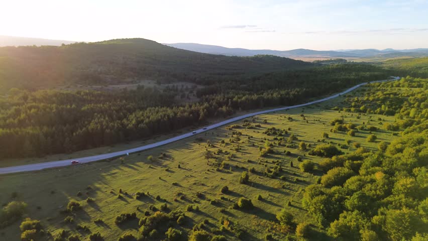 Aerial view of cars and trucks driving along a highway through a green forest and hills at sunset