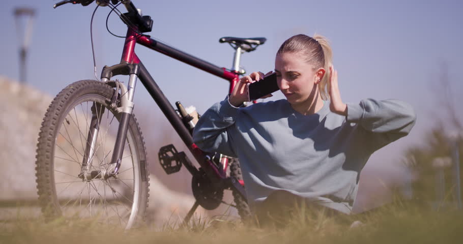Active young female cyclist pauses in a grassy field to talk on her mobile phone, with her mountain bike beside her