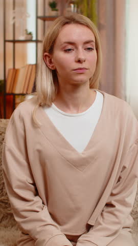 Young adult woman holds cheek in pain while sitting on home sofa, showing signs of strong toothache. Blonde girl frowns and shows discomfort, reacting emotionally to dental problem and sensitivity