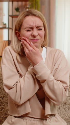 Young adult woman holds cheek in pain while sitting on home sofa, showing signs of strong toothache. Blonde girl frowns and shows discomfort, reacting emotionally to dental problem and sensitivity