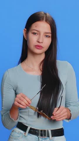 Caucasian young stylish cool woman wearing yellow sunglasses, charming friendly playful smile, celebrate victory win. Teenager happy girl feel satisfied excitement isolated on blue background indoors