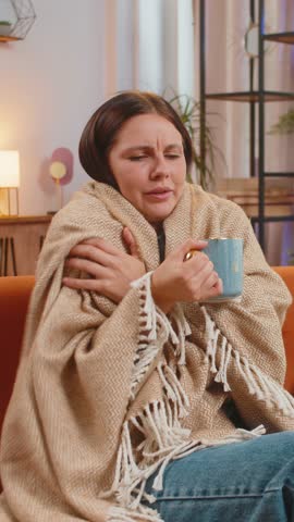 Young woman covered by warm blanket drinks coffee at home. Caucasian female freezes from the cold, feels bad, unhealthy. Sick girl with flu symptoms covered with blanket warms up with hot tea at home.
