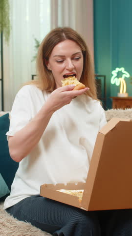 Pregnant middle-aged woman at home opens pizza box smells slice bites smiles tastes dinner treat. Caucasian girl on sofa enjoys warm cheesy food big full comfort feeds hunger thinks baby moves inside.