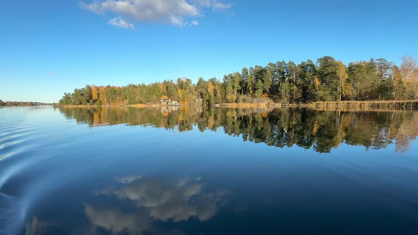 Autumn baltic sea view in nordic archipelgago. Forest with lush green and yellow trees surround the shore. Wide shot with clouds reflected on the water surface. Shot moves slowly and steady. Serene. 