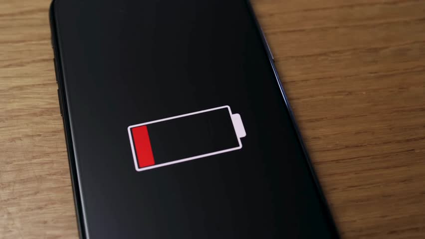Smartphone screen displays low battery icon, emphasizing urgency. Soft light ambient in background suggests evening time. Device resting on wooden surface, highlighting modern technology reliance.