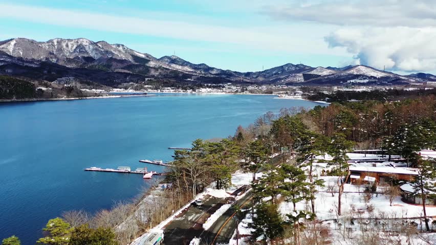 Clear blue water reflects majestic mountains under bright sky. Pine trees line shores, creating peaceful atmosphere in winter setting. Ideal for nature enthusiasts and travel content.