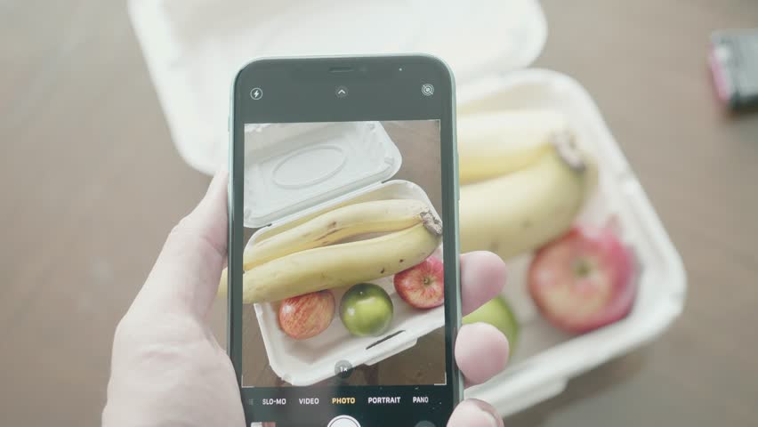A hand holds a smartphone, photographing fresh fruits including bananas, apples, and a lime, arranged on a table. Perfect for showcasing healthy eating and creative food photography.