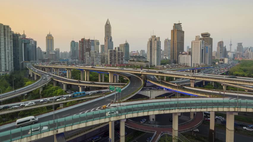 Dynamic aerial shot captures a vibrant interchange with multiple highways and buildings, showcasing the energy of city travel during sunset hours.