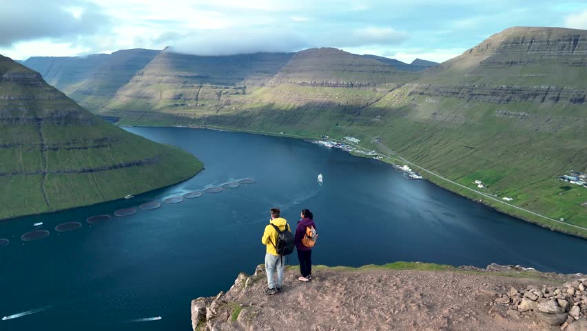 Two hikers enjoy a stunning landscape in Klakkur Faroe Islands, gazing at the fjord and majestic mountains. The tranquil waters reflect the sky while verdant hills rise dramatically around them.