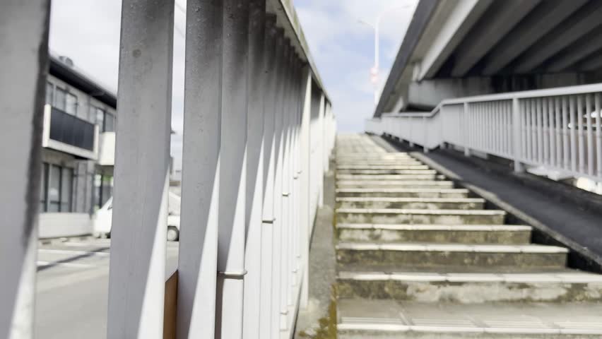 A low-angle view of a pedestrian overpass stairway with white metal railings and concrete steps under a bright cloudy sky in a quiet urban area.