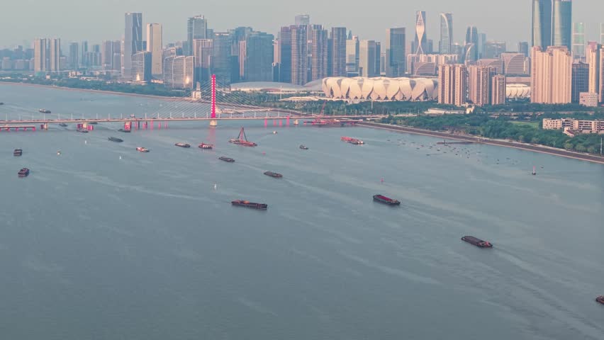 Cargo ships and boats navigate the Qiantang River in Hangzhou China. A red bridge spans the river, with the city skyline visible in the distance