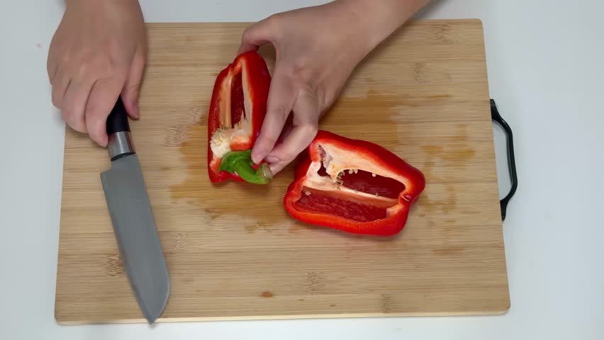 Woman uses a knife to remove seeds from a red pepper on a wooden cutting board at a kitchen table