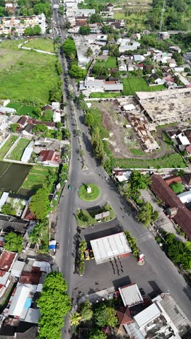 Aerial drone view of a town with a traffic roundabout intersection, gas station, and surrounding houses and green fields.
