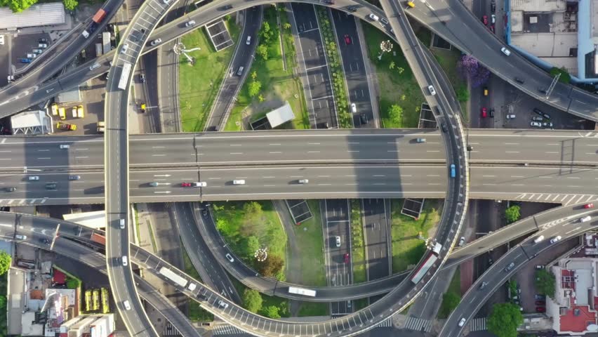 Aerial perspective of a busy city highway interchange showcases vehicles navigating roundabouts and green areas amidst urban architecture.