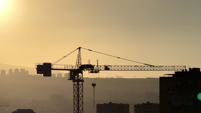 Construction crane against the backdrop of a city with a bright sun . A crane is in the sky above a city at day The crane is tall and is in the middle of the sky