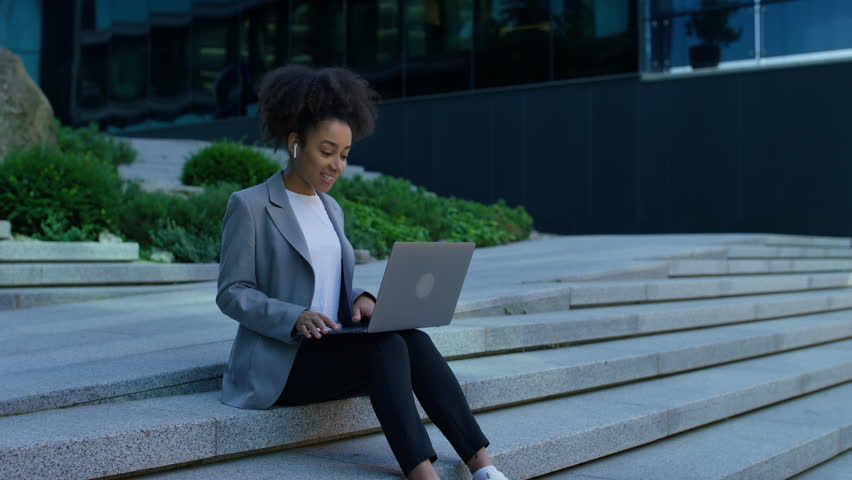 Professional young woman in blazer and earbuds sitting on city steps, gesturing while engaged in a video call on her laptop.