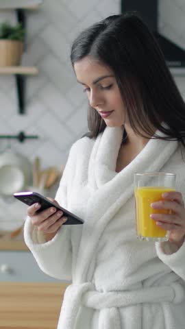 A woman in a bathrobe stands in her kitchen, smiling as she checks her smartphone and holds a glass of orange juice.