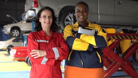 Two professional female mechanics Caucasian woman in red and  african american woman in yellow holding a tablet stand side-by-side and confidently give thumbs-up signs in a car garage - Powered by Shutterstock - Get 15% off with code: PIKWIZARD15