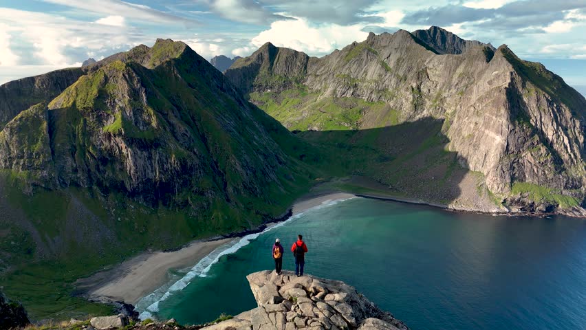 Hikers admire the stunning landscape from the summit of Ryten. The pristine beach of Kvalvika lies below, framed by majestic mountains. A perfect day for exploring Lofotens natural beauty.