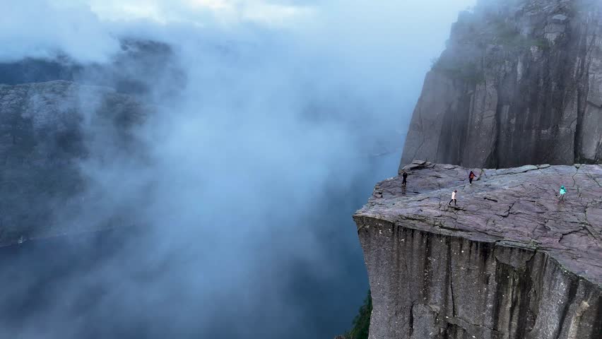 Hikers explore the breathtaking Preikestolen, a famous cliff in Norway that offers stunning views of the fjord below. The scene captures the dramatic cliffs shrouded in mist and clouds.