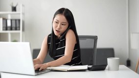 Busy young asian businesswoman having phone call conversation while multitasking, typing on laptop computer and writing down notes in a notebook at her modern office desk - Powered by Shutterstock - Get 15% off with code: PIKWIZARD15