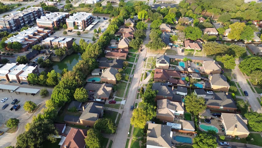 Modern white-roof apartment complex with pond, pool, covered parking, surrounded by mature trees, upscale homes with backyard pools, showcasing integrated housing density in Denton County, Texas. USA