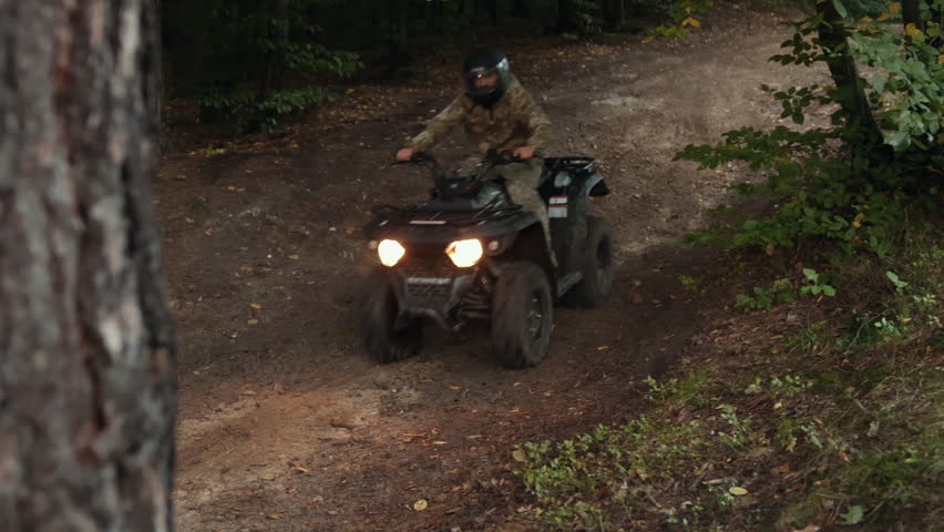 Camouflage clothing, Quad driver, Dense woods. In dense forest, ATV rider wearing helmet and camouflage clothing navigates dirt trail with headlights on.