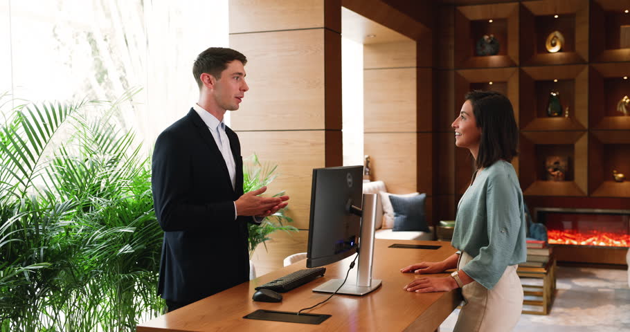 Young 30s Hispanic male receptionist in formal attire welcomes guest at reception desk, talking, providing check-in services and answering questions in modern hotel lobby with elegant interior design