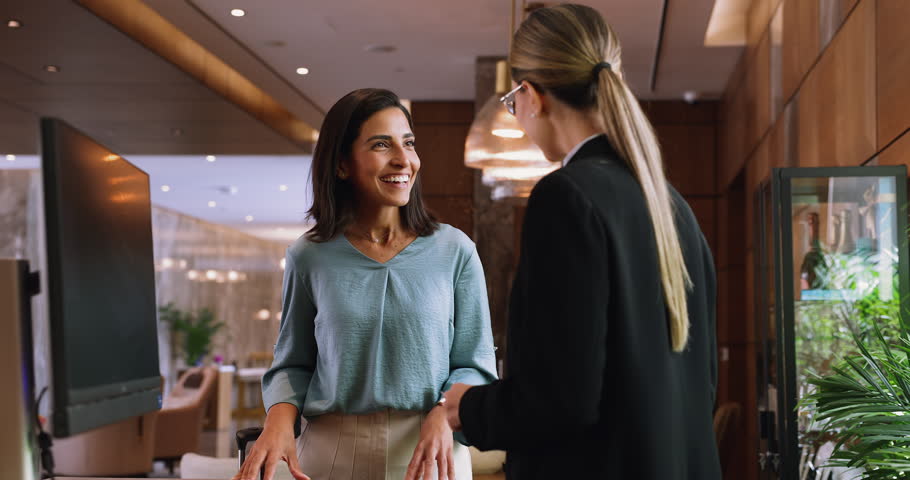 Friendly female hotel receptionist hands room keycard to woman guest at front desk, guest has just arrived with luggage, smiling, receiving keys. Travel, tourism, customer services, hospitality, trip
