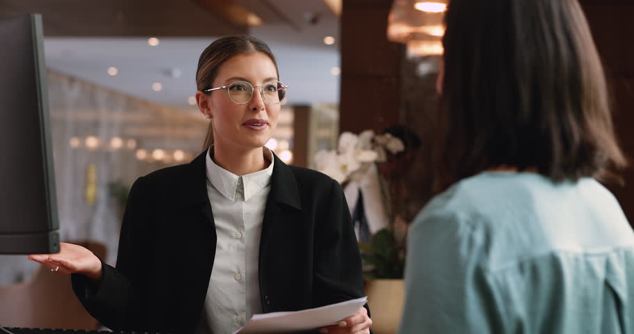 Smiling Hispanic woman receptionist in elegant suit holding papers with guest registration form, asking or provide information, talking with guest at front counter desk in modern boutique hotel lobby