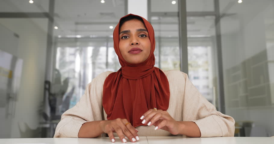 Positive young Indian business woman in Muslim hijab waving hand hello at camera, smiling, talking on online video conference call, sitting at office table, listening, nodding