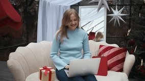 Cheerful woman sitting on cozy sofa with laptop, waving hand and smiling while talking on video call. Christmas decorations and gift box nearby. Concept of online communication, holiday greetings - Powered by Shutterstock - Get 15% off with code: PIKWIZARD15