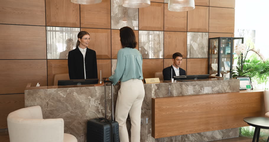 Friendly female hotel receptionist in elegant formal attire hands keycard to Brazilian woman guest at front desk during check-in. Smiling guest with luggage, receives room key. Business trip, travel