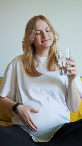 Video of pregnant woman sitting on bed, holding her belly and drinking water. Cozy home atmosphere with soft light. Concept of healthy lifestyle during pregnancy, hydration, self-care, and motherhood.