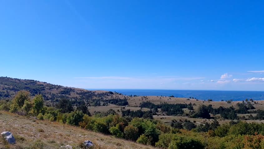 Shot capturing the vast Učka Nature Park, Croatia. A serene, sunny mountain landscape of rolling green hills and scattered trees, with the distant blue Adriatic Sea on the horizon under a clouds