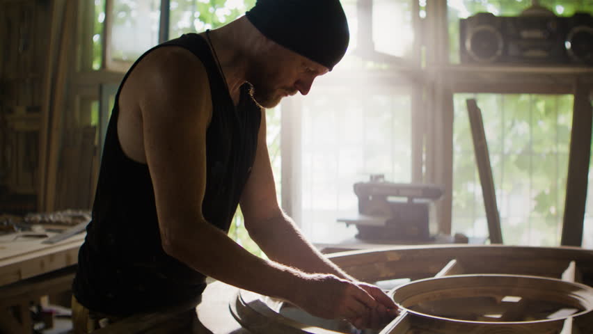 Carpenter in hat restores round wooden frame with paste in workshop. Skilled craftsman repairs small flaws of vintage furniture with manual tools in shop