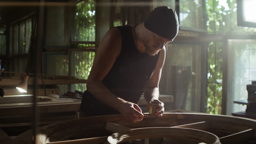 Young man in hat covers flaws with lacquer onto round window frame in carpentry. Focused bearded craftsman restores antique door with material in craft shop