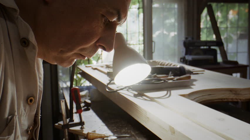 Skilled man bends over wooden door blank with focused face. Artisan moves carving tool slowly adjusting line depth with precision sitting near bright lamp