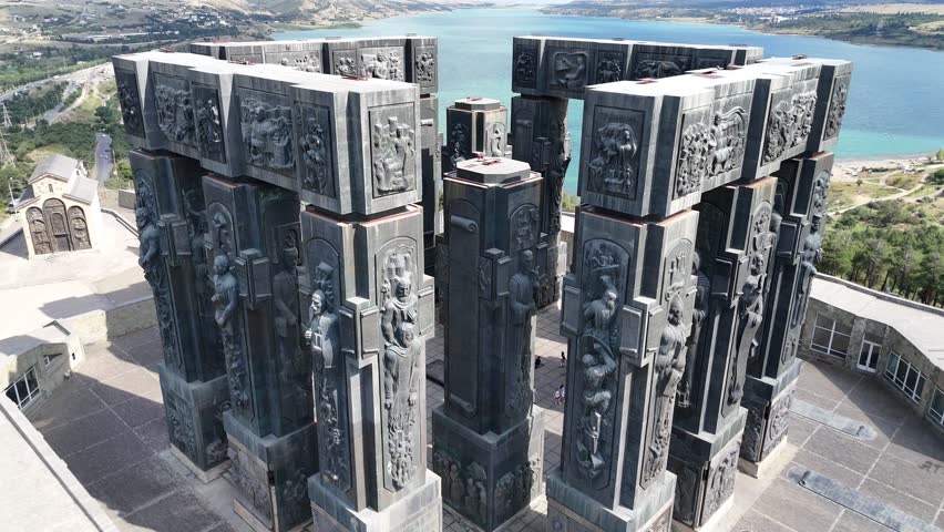 Aerial view of the Chronicle of Georgia Monument with tall stone pillars depicting Georgian kings and Christian history, with the beautiful Tbilisi Sea in the background.