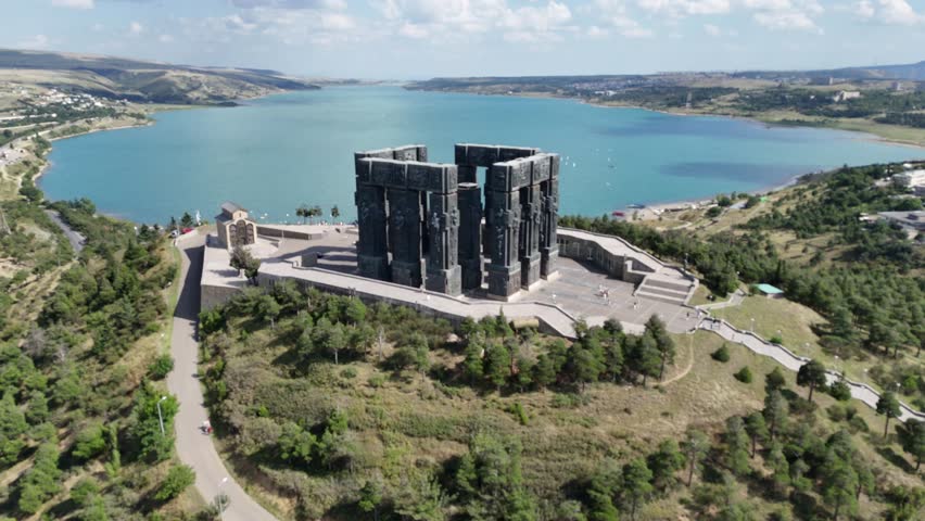Aerial view of the Chronicle of Georgia Monument with tall stone pillars depicting Georgian kings and Christian history, with the beautiful Tbilisi Sea in the background.