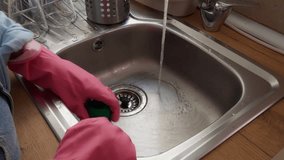 Female hands in pink rubber gloves scrubbing stainless steel kitchen sink with green brush under running water, illustrating daily household cleaning and hygiene routine - Powered by Shutterstock - Get 15% off with code: PIKWIZARD15