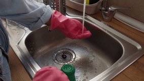Woman's hands in protective pink rubber gloves use green brush to wash and clean stainless steel kitchen sink with running water, demonstrating daily household chores and hygiene - Powered by Shutterstock - Get 15% off with code: PIKWIZARD15