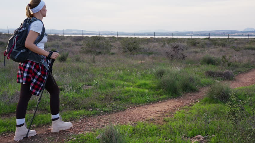 Young woman traveler with backpack and hiking poles strolling along dusty trail in lush field, embracing spirited outdoor existence while taking in beauty of scenery